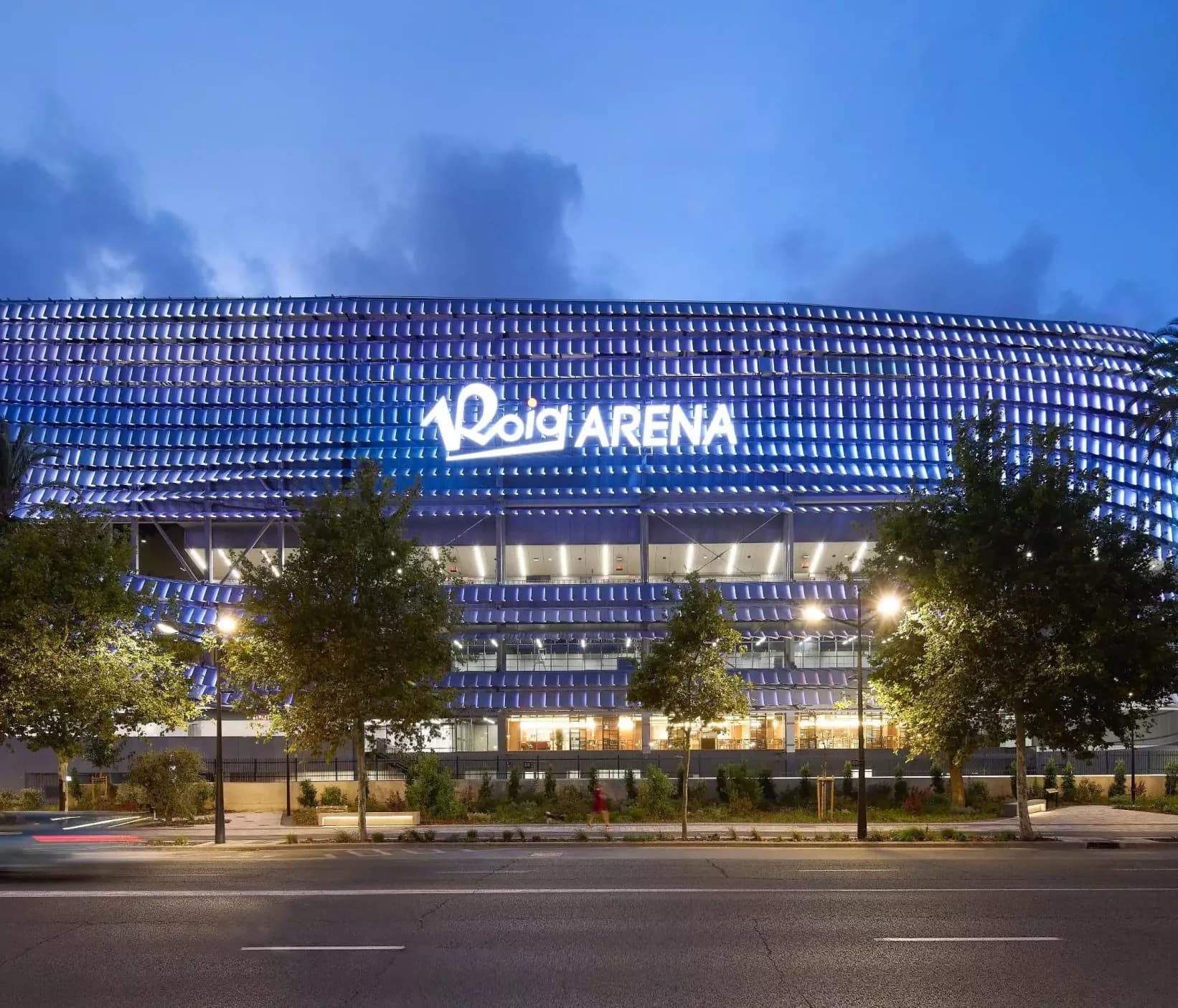 Vista frontal del Roig Arena de noche con la fachada iluminada en azul.